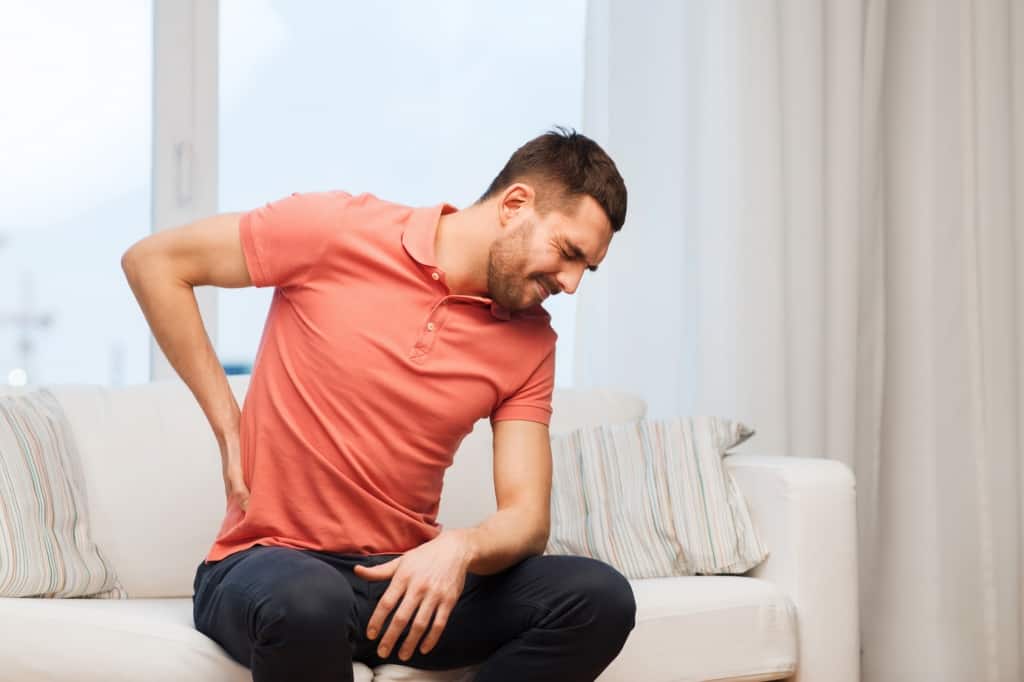man sitting on white couch in salmon shirt rubbing his back while in pain - first chiropractic visit